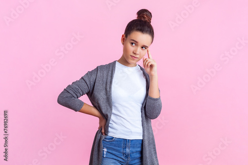 Portrait of concentrated pensive teenage girl with bun hairstyle in casual clothes standing keeps finger on temples thinking about university. Indoor studio shot isolated on pink background.