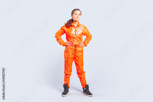 Full length portrait of confident hip-hop dancer teenage girl with brunette hair wearing bright orange jumpsuit standing with hands on waist. Indoor studio shot isolated on gray background. © khosrork