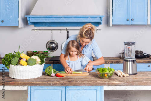 Blonde woman and little female child cooking healthy salad together, cutting chopping vegetables, preparing vegetarian food in modern kitchen, mother teaching daughter to make dishes. photo