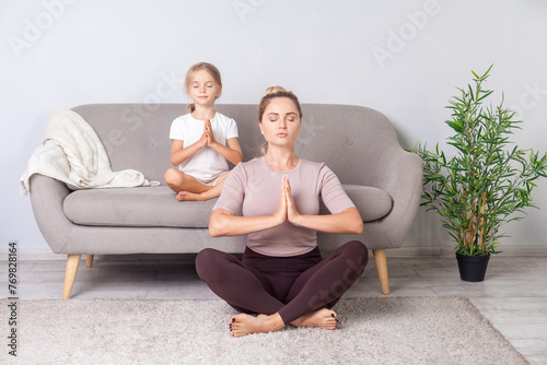 Mindfulness and harmony. Lotus posture. Calm woman with her daughter with closed eyes and prayer gesture practicing yoga together, doing exercise breath technique, meditating in room sitting on floor.