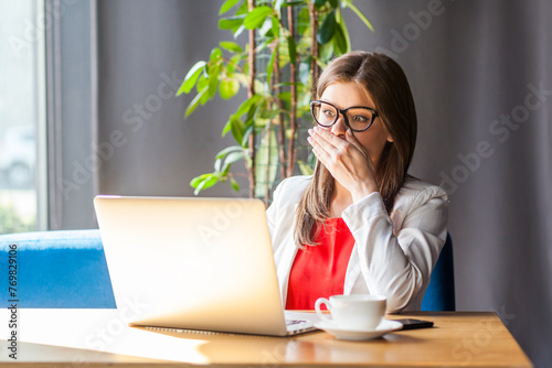 Portrait of shocked scared woman in glasses working on laptop sees something awful on display has no words, wearing jacket and re shirt. Indoor shot, cafe or office background. photo