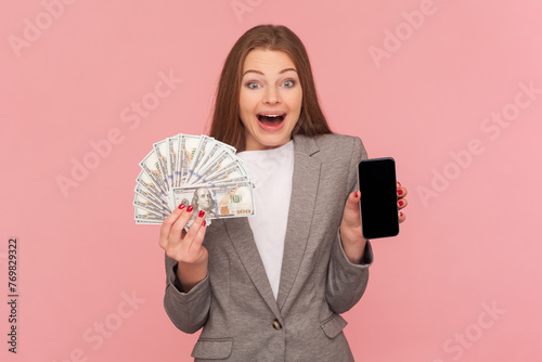 Portrait of amazed surpised woman with brown hair holding dollar banknotes and smart phone with empty display, wearing business suit. Indoor studio shot isolated on pink background.