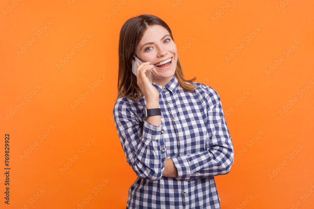 Portrait of happy smiling cheerful woman with brown hair standing talking on cell phone hearing pleasant news, wearing checkered shirt. Indoor studio shot isolated on orange background Portrait of happy smiling cheerful woman with brown hair standing talking on cell phone hearing pleasant news, wearing checkered shirt. Indoor studio shot isolated on orange background