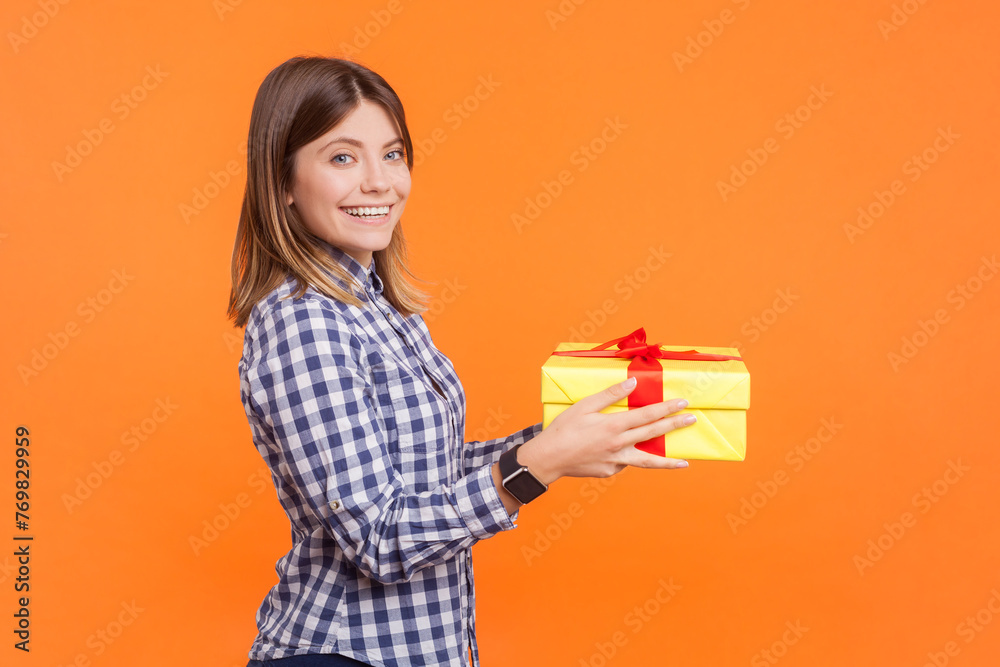 Profile portrait of smiling happy optimistic woman with brown hair giving present to boyfriend looking at camera, wearing checkered shirt. Indoor studio shot isolated on orange background Profile portrait of smiling happy optimistic woman with brown hair giving present to boyfriend looking at camera, wearing checkered shirt. Indoor studio shot isolated on orange background