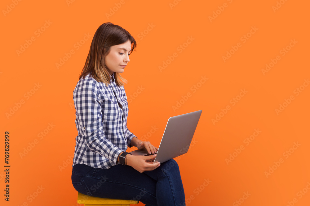 Side view portrait of confident woman with brown hair typing on laptop keyboard, working remotely, wearing checkered shirt. Indoor studio shot isolated on orange background Side view portrait of confident woman with brown hair typing on laptop keyboard, working remotely, wearing checkered shirt. Indoor studio shot isolated on orange background