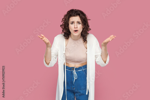 Portrait of angry beautiful young adult woman with curly hair wearing casual style outfit raised her hands, asking why, arguing with somebody. Indoor studio shot isolated on pink background.