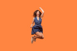 Full length portrait of extremely happy brunette woman wearing denim dress jumping up high with clenched fists celebrating her success. Indoor studio shot isolated on orange background.