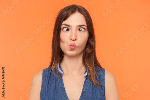 Portrait of childish funny crazy brunette woman wearing denim dress crossing her eyes making fish lips having fun grimacing. Indoor studio shot isolated on orange background.