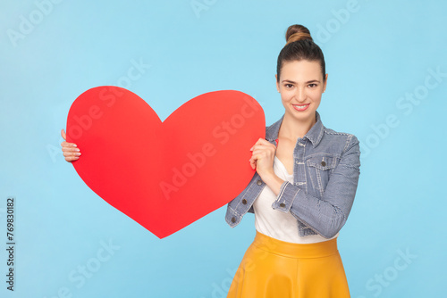 Portrait of romantic smiling woman with bun hairstyle wearing denim jacket holding big read heart saying about her feelings. Indoor studio shot isolated on light blue background.