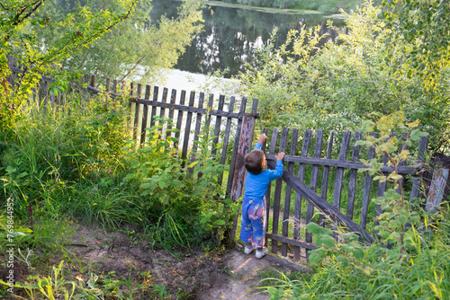 boy standing in care, child in the village, life outside the city, old gate, picket fence, lake, childhood, baby waiting by the fence