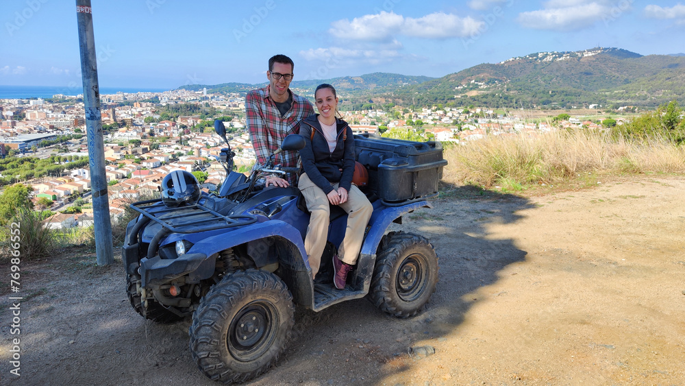 Fototapeta premium Young couple on the top of the mountain with the quad, off-road route through the mountain to the viewpoint of the coastal town. They are posing for the photo with the off-road vehicle.