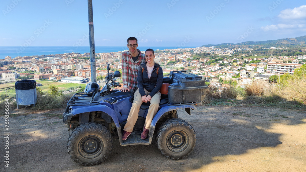 Fototapeta premium Young couple on the top of the mountain with the quad, off-road route through the mountain to the viewpoint of the coastal town. They are posing for the photo with the off-road vehicle. 