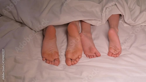 restless legs of the boy son child and the mother female foot lie under the blanket on the sheet in the evening. Baby kid girl daughter and woman mom wiggling bare feet in bed in the bedroom photo