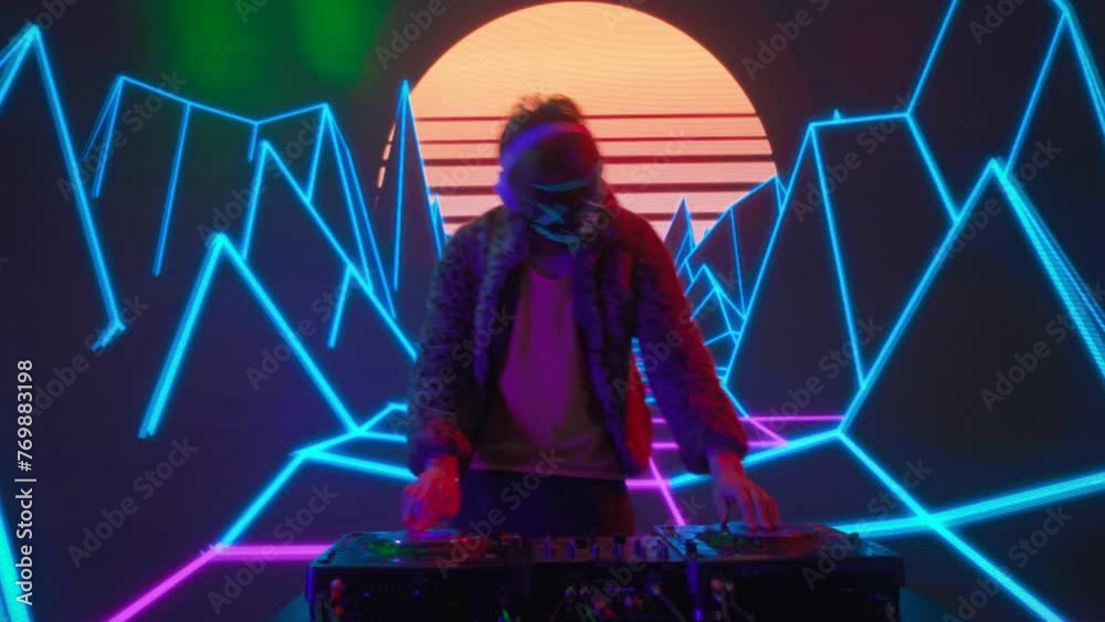 Attractive male on digital wall colorful visual graphic background. Young man dj in mask dancing, mixing plays music on turntables mixer at night club.
