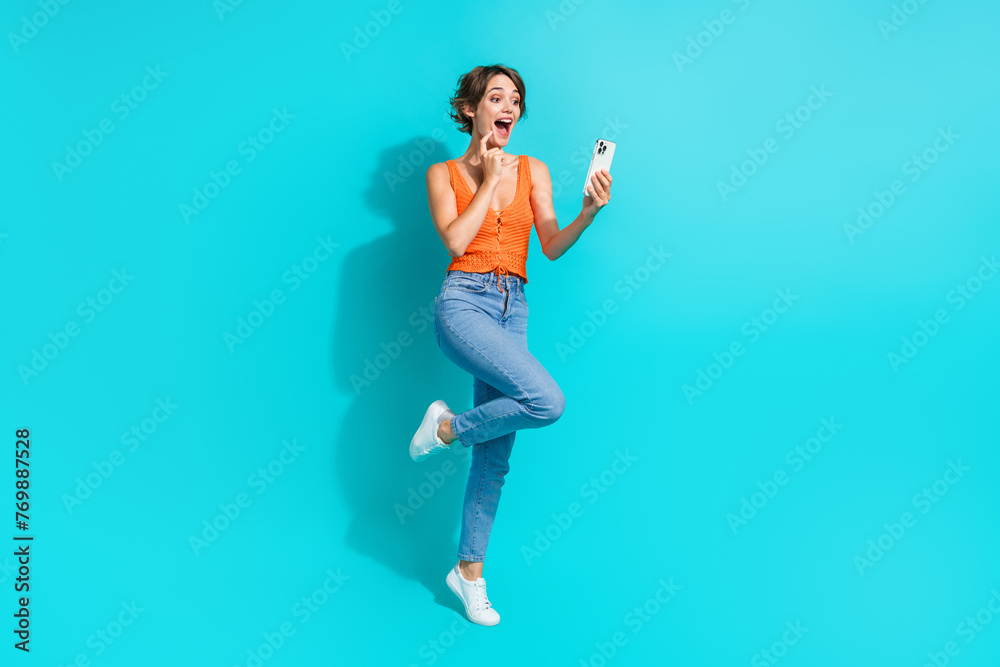 Full length photo of overjoyed funky woman wear orange singlet jeans look at eshop in smartphone isolated on turquoise color background