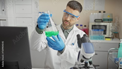 A focused man conducts experiments in a laboratory wearing protective glasses and gloves while examining a green liquid. photo