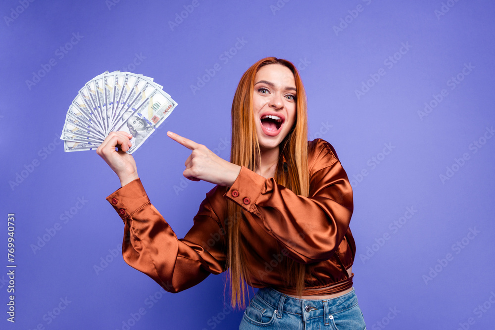 Portrait of nice young woman indicate finger dollar bills wear brown shirt isolated on purple color background Portrait of nice young woman indicate finger dollar bills wear brown shirt isolated on purple color background
