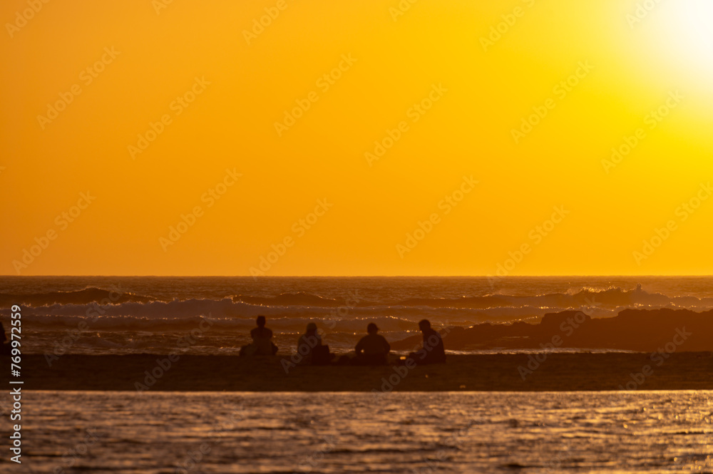 Group of men and women sitting and looking at sunset on ocean beach, orange sky, silhouettes of people on vacation