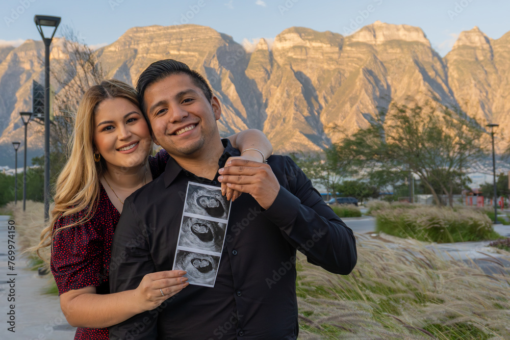 Couple Embracing with Ultrasound Photos in Front of Mountains, latin young couple