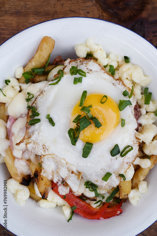 Top view of fried potatoes with melted cheese, chorizo, bell pepper, onion and a grilled egg in a white bowl on the wooden table.	