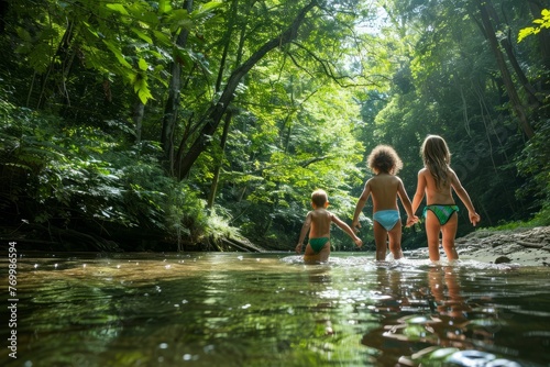 Three children in bathing suits are wading in a river against a forest backdrop in a low angle shot