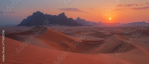   The sun descends on a desert panorama featuring sandy dunes upfront and a distant rock formation photo