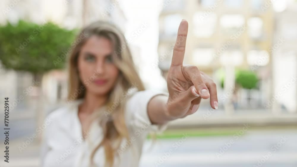 Cool, young blonde woman exudes relaxed, yet serious energy, standing outdoors in sunny, urban street, resolutely saying no with her finger while maintaining eye contact with camera Cool, young blonde woman exudes relaxed, yet serious energy, standing outdoors in sunny, urban street, resolutely saying no with her finger while maintaining eye contact with camera