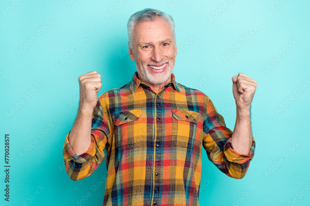 Photo of positive lucky man pensioner dressed checkered shirt smiling rising fists isolated teal color background