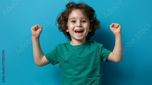 Cheerful young child in green shirt raising fists in victory pose. Studio portrait against blue background photo