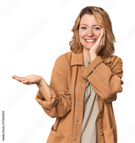 Blonde middle-aged Caucasian woman in studio holds copy space on a palm, keep hand over cheek. Amazed and delighted.