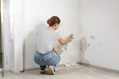 Saltpeter on the wall problem. Woman is using a scraper to scrape and remove all loose paint and plaster that is in poor condition, until a firm surface is achieved. 