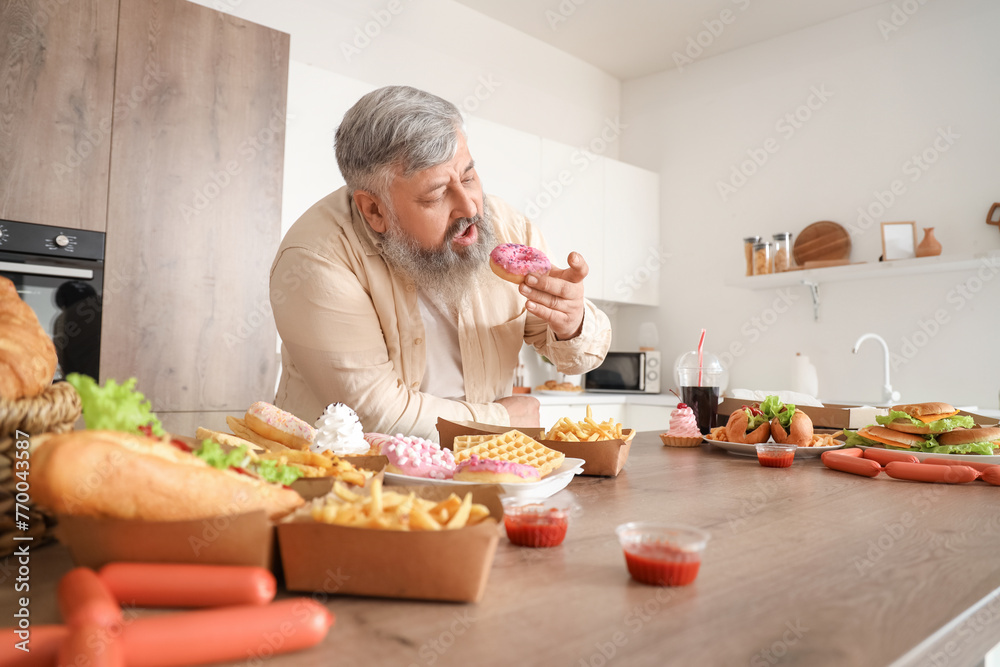 Overweight mature man at table full of unhealthy food in kitchen. Overeating concept Overweight mature man at table full of unhealthy food in kitchen. Overeating concept