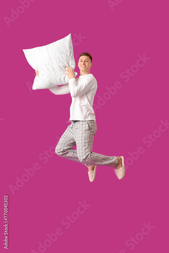 Young man in pajamas with pillow jumping on purple background
