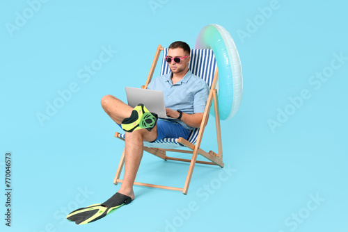 Young office worker in flippers with inflatable ring and laptop sitting on chaise lounge against blue background