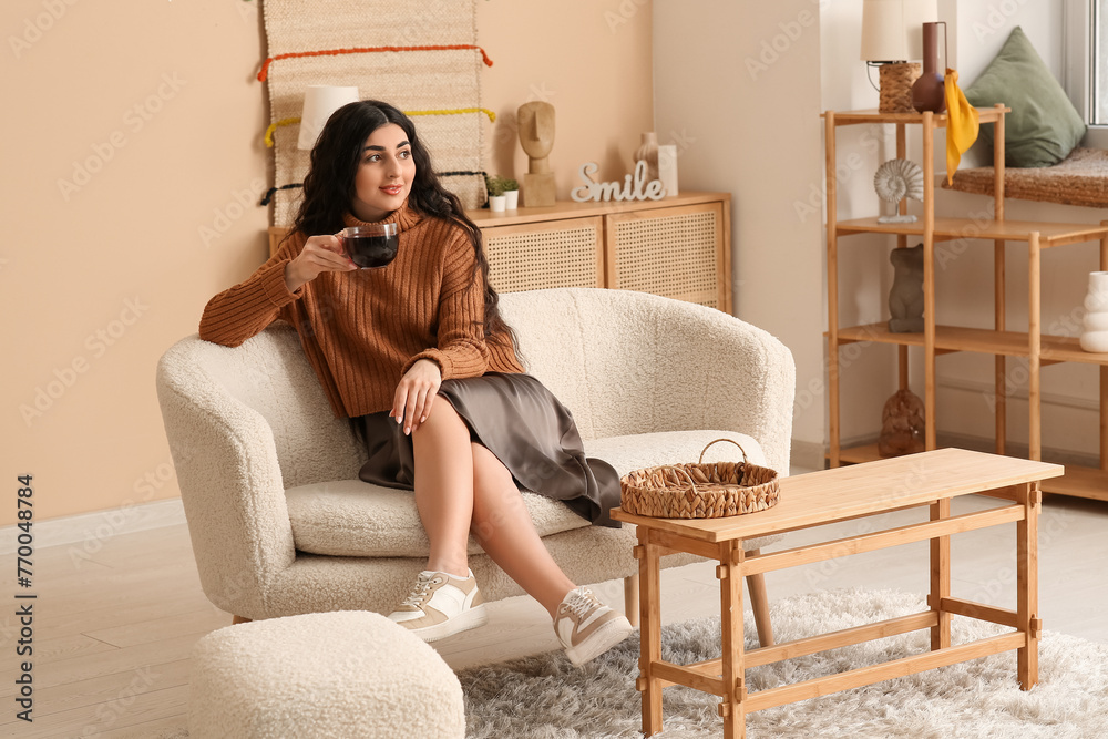 Obraz premium Beautiful young woman with glass cup of hot tea on sofa in living room