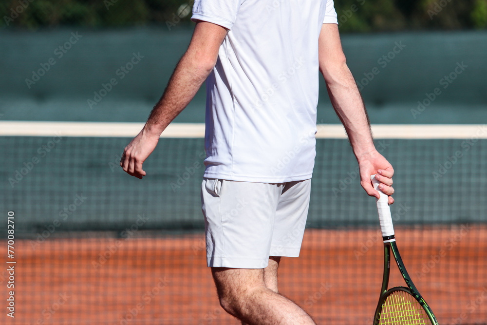 Close up of young boy with racket playing tennis on a clay court during a university tournament. The tennis player is wearing a white sports suit and is intent on serving the set point