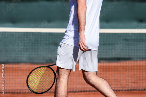 Close up of young boy with racket playing tennis on a clay court during a university tournament. The tennis player is wearing a white sports suit and is intent on serving the set point © Vamos Sports Prod