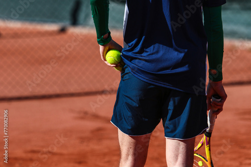 Detail of young boy with racket playing tennis on a clay court during a university tournament. the athlete is wearing a blue sports t-shirt and is preparing to serve for set point