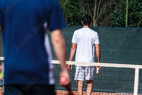 Close up of young boy with racket playing tennis on a clay court during a university tournament. The tennis player is wearing a white sports suit and is intent on serving the set point