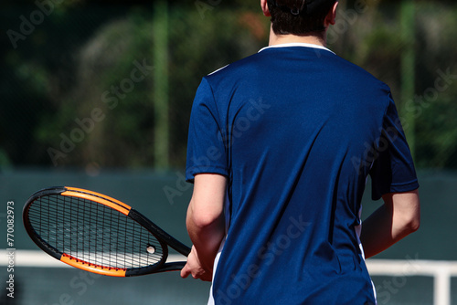 Close up of young boy with racket playing tennis on a clay court during a university tournament. the athlete is wearing a blue sports shirt.