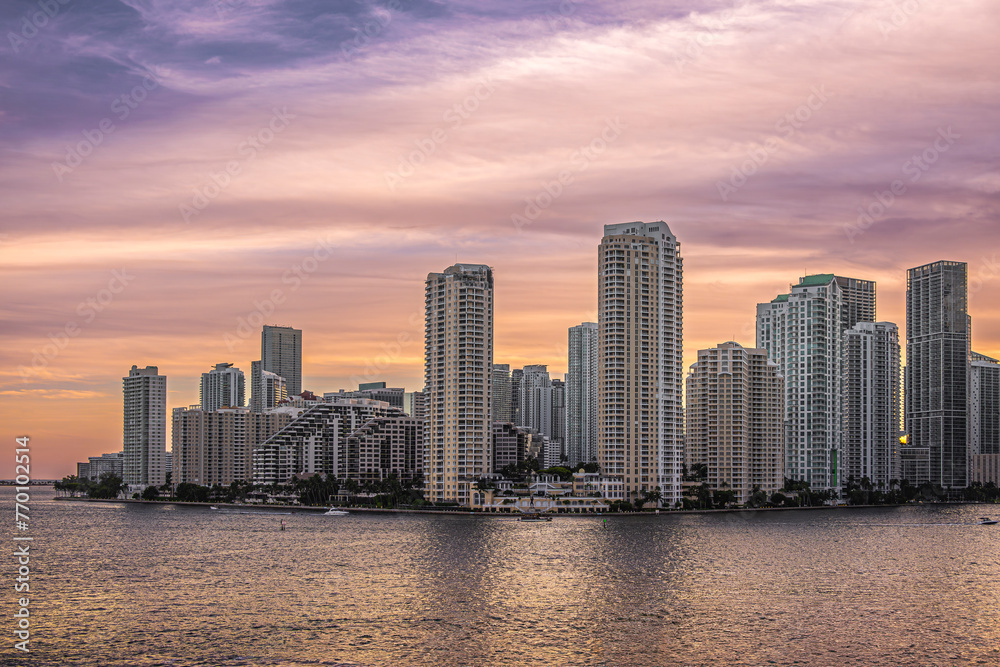 Miami, Florida, USA - July 29, 2023: Warm colored sky over buildings on Brickell Key island at evening 19:43. The Centinel statue between Tequesta points on the shoreline. Green foliage