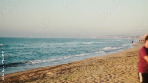 Calm waves. People walk on sandy beach at dawn in summer. Birds and seagulls fly. City, mountains on the horizon. Atmospheric video, for shooting retro. Shooting from a tripod. Algae coast. Day photo