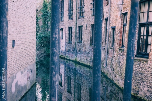Vintage-toned image of a serene canal with reflections of old brick buildings, viewed through iron bars, evoking a sense of history and tranquility.