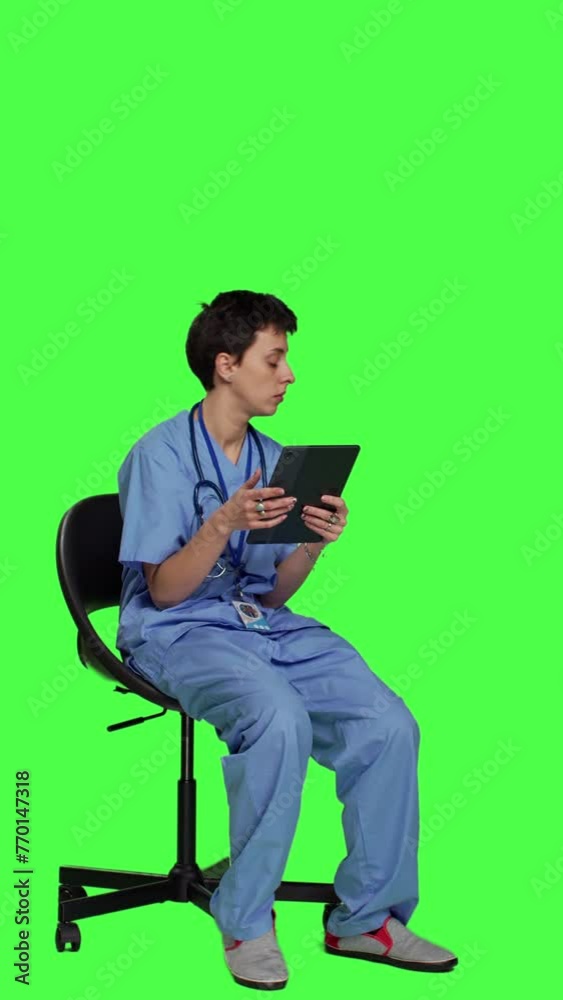 Side view Medical assistant browsing internet on tablet while she sits down in a chair, killing time by scrolling on social media apps and texting friends. Nurse in scrubs using modern gadget. Camera Side view Medical assistant browsing internet on tablet while she sits down in a chair, killing time by scrolling on social media apps and texting friends. Nurse in scrubs using modern gadget. Camera