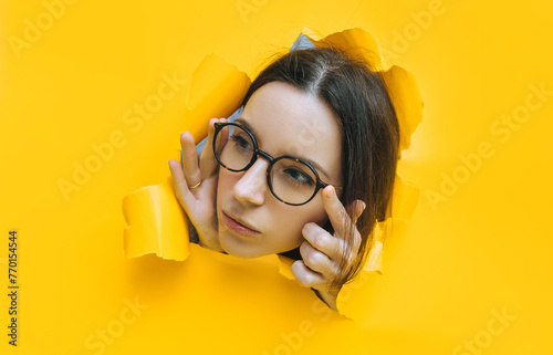 Portrait of a cute young woman with glasses looking through a hole in yellow paper. An incredulous look. Women's curiosity and gossip. Jealous wife.