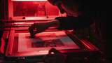 In a 1960s-style darkroom, a photographer adjusts an enlarger projecting a black and white image onto Ilford paper, under the glow of red safety lights.