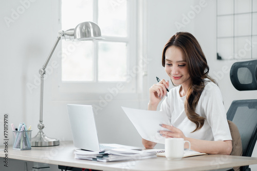 Smiling businesswoman in a bright office reviewing documents with a pen in hand, showing efficiency and focus in her professional routine. © Mongta Studio