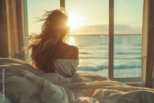 Woman on bed looking out the window overlooking the sea and sunrise #770201516
