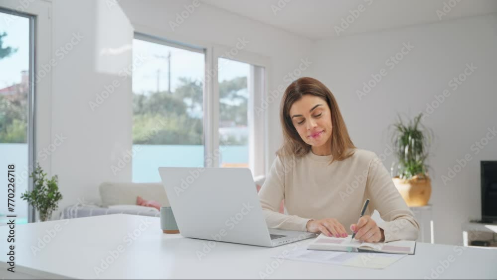 Happy middle aged woman using laptop computer sitting at home table elearning. Smiling older mature lady hybrid working, watching webinar at home, typing, browsing writing notes in living room.