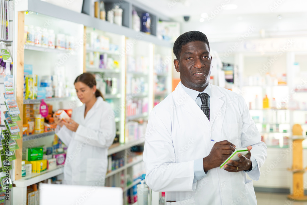 Male pharmacist standing with notebook in salesroom of drugstore. His colleague standing behind.